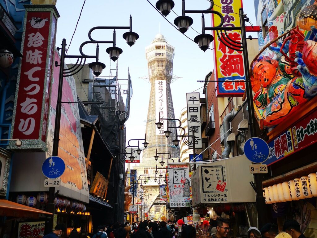 Eine belebte Straße in Osaka, Japan, voller Menschen, farbenfroher Ladenschilder und dekorativer Laternen, mit dem ikonischen Tsūtenkaku-Turm im Hintergrund, der sich unter einem klaren Himmel der Kansai region erhebt.