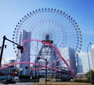 Ein großes Riesenrad mit einer Digitaluhr in der Mitte steht vor dem blauen Himmel von Yokohama, umgeben von hohen modernen Gebäuden und einer rosafarbenen Achterbahnstrecke, die sich vor ihm windet.