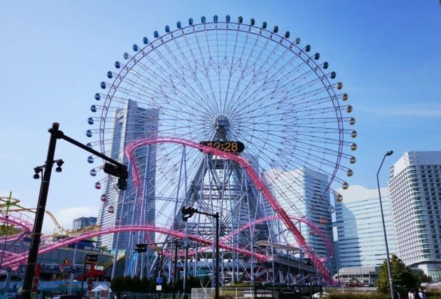 Ein großes Riesenrad mit einer Digitaluhr in der Mitte steht vor dem blauen Himmel von Yokohama, umgeben von hohen modernen Gebäuden und einer rosafarbenen Achterbahnstrecke, die sich vor ihm windet.