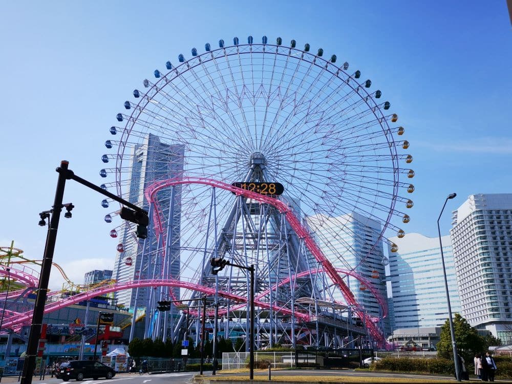 Ein großes Riesenrad mit einer Digitaluhr in der Mitte steht vor dem blauen Himmel von Yokohama, umgeben von hohen modernen Gebäuden und einer rosafarbenen Achterbahnstrecke, die sich vor ihm windet.
