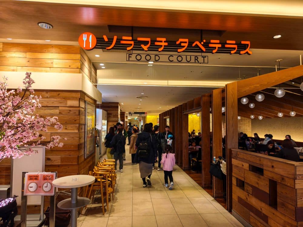 Eingang zu einem Food Court im Skytree, mit einem warmen Holzinterieur, Kirschblütendekor auf der linken Seite und einem Schild auf Japanisch mit der Aufschrift ソラマチタベテラス mit FOOD COURT darunter, während mehrere Personen eintreten.