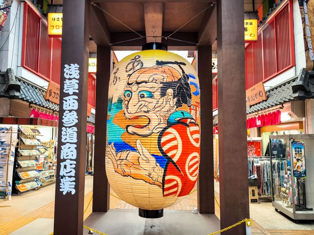 Eine große, farbenfrohe japanische Papierlaterne mit einer traditionellen Illustration hängt in der überdachten Einkaufsstraße von Asakusa. Die umliegenden Geschäfte bieten verschiedene Waren an, während Schilder mit japanischen Schriftzeichen die kulturelle Atmosphäre des Viertels unterstreichen.