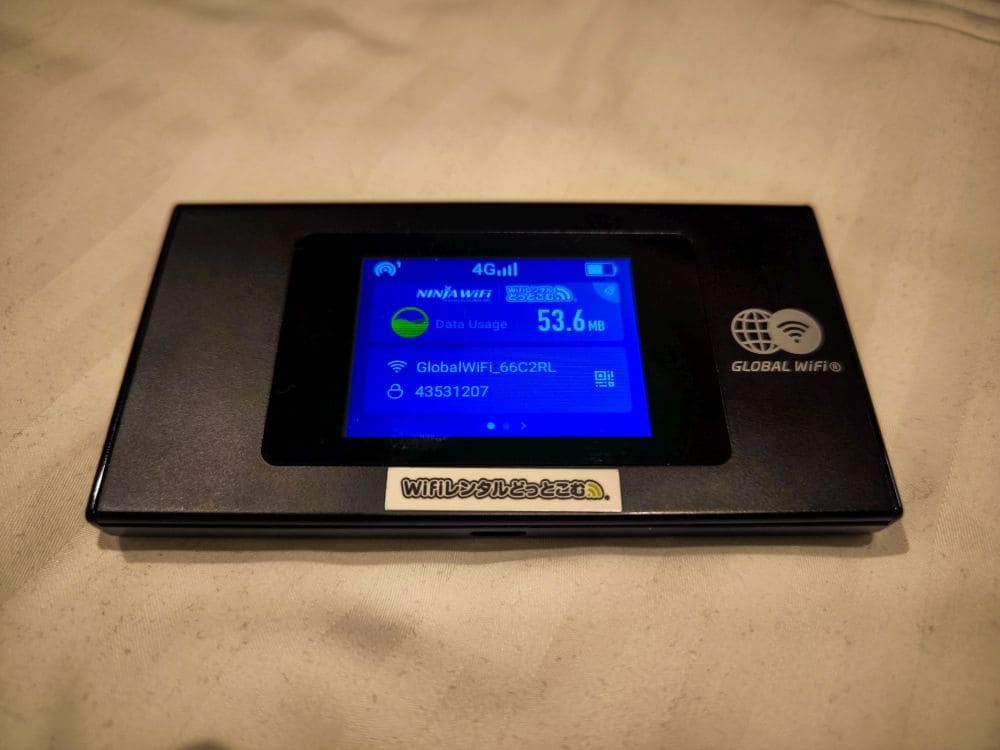 Das elegante, schwarze Ninja WiFi-Gerät verfügt über eine digitale Anzeige, die 53,6 MB Datennutzung und Netzwerkdetails anzeigt. Das tragbare Gerät mit seinem klaren Bildschirm und den Tasten liegt elegant auf einer hellen Stoffoberfläche.
