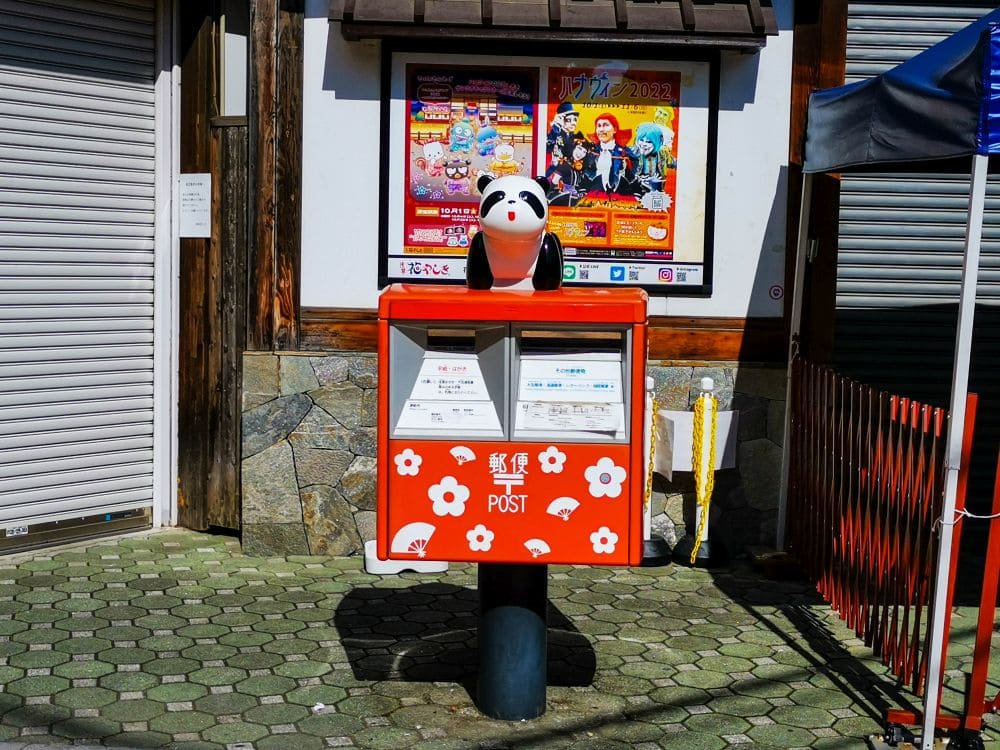 Ein roter Briefkasten, geschmückt mit weißen Blumen und einer Pandafigur darauf, steht stolz auf einem Steinweg in Asakusa. Dahinter schmücken bunte Plakate und geschlossene Fensterläden die Hauswand und fangen den lebendigen Geist dieses ikonischen Viertels ein.