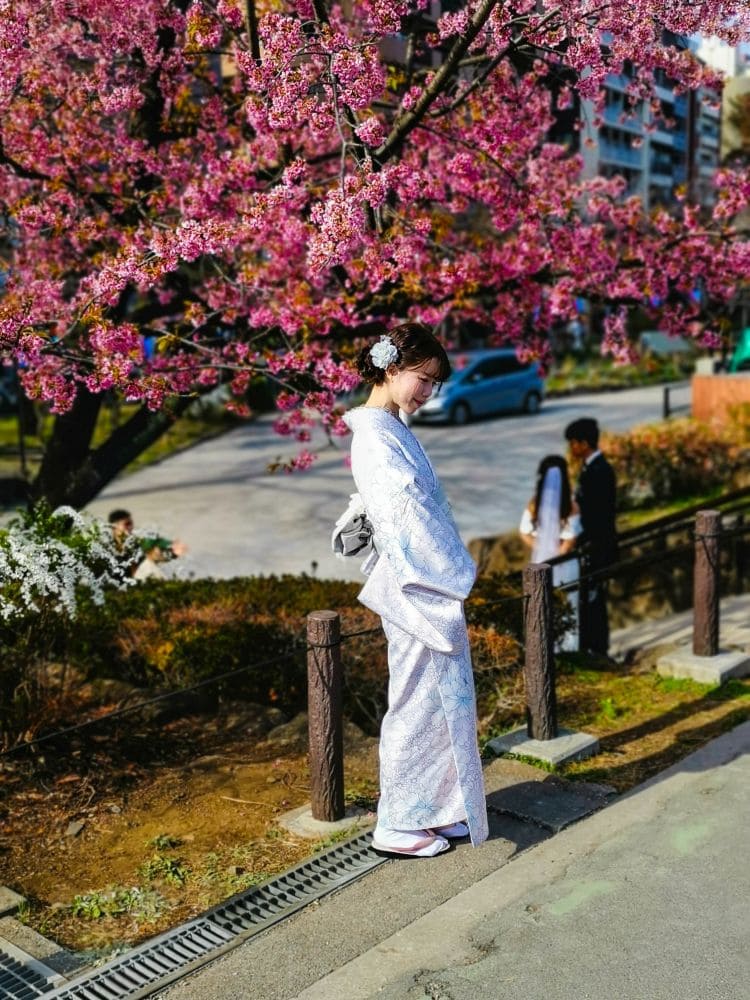 Eine Frau im traditionellen Kimono steht an einem Weg in Asakusa, über ihr leuchtend rosa Kirschblüten. Sie blickt nach unten, Blumen schmücken ihr Haar. Im Hintergrund sind Menschen und Gebäude zu sehen, die eine ruhige Frühlingsszene einfangen.