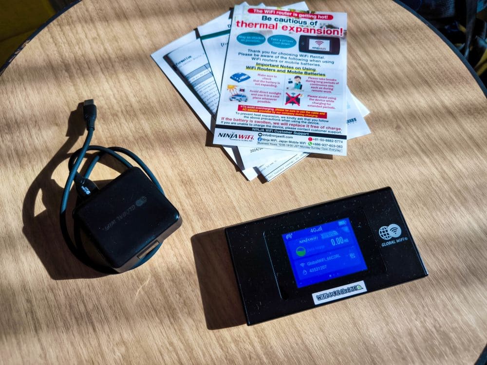 Ein Gerät mit Bildschirm und Ladekabel liegt auf einem Holztisch, neben einer Anleitung für Ninja Wifi. Daneben liegt eine bunte Broschüre zur Wärmeausdehnung. Sonnenlicht wirft Schatten auf den Tisch und verleiht der Szene eine ruhige Aura.