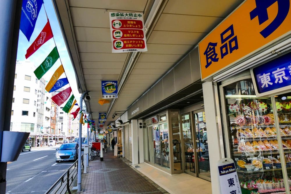 Eine belebte Straße in Tokio, die an die Kappabashi-Straße erinnert, ist mit bunten Fahnen geschmückt, die von einem überdachten Gehweg hängen. Geschäfte locken Küchenliebhaber mit ihren Schaufenstern. Darüber hängen Schilder mit japanischem Text, während am Straßenrand ein blaues Auto parkt.