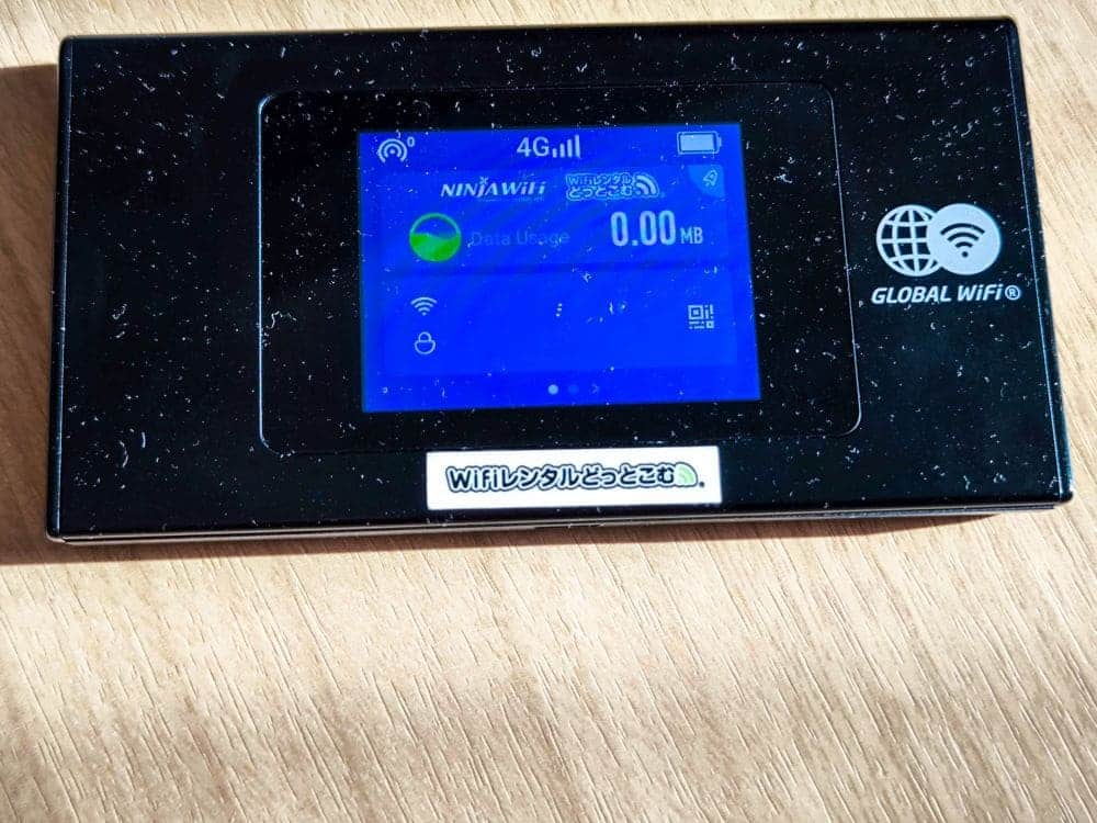 Ein schwarzes tragbares WLAN-Gerät liegt auf einer Holzoberfläche. Auf dem Display wird unter dem bekannten „Ninja WiFi“ die Signalstärke „4G“ und ein Datenverbrauch von 0,00 MB angezeigt. Rechts steht „GLOBAL WiFi“, unter dem Display befindet sich eine japanische Beschriftung.
