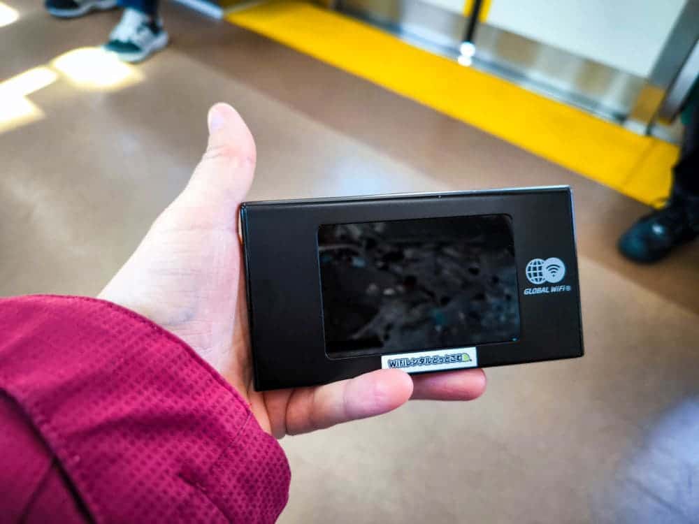Eine Nahaufnahme zeigt eine Person, die ein elegantes Ninja-WLAN-Gerät in einer leuchtend roten Hülle in der Hand hält. Der Innenraum ist mit einem braunen Boden und einer gelben Sicherheitsleine ausgestattet, was an die Hektik eines Verkehrsknotenpunkts erinnert.