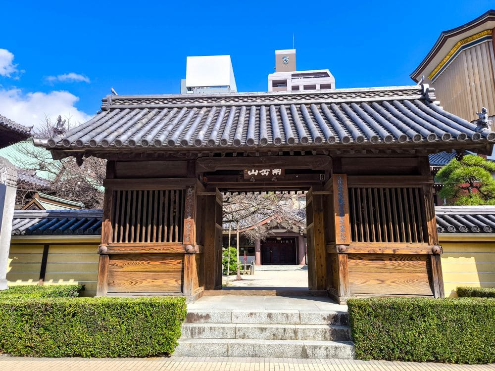 Ein traditionelles japanisches Holztor mit Ziegeldach im Tochoji-Tempel führt zu einem ruhigen Innenhof. Die umliegenden Gebäude bilden einen wunderschönen Kontrast zum klaren blauen Himmel, während gepflegte Hecken den Weg einladend säumen.