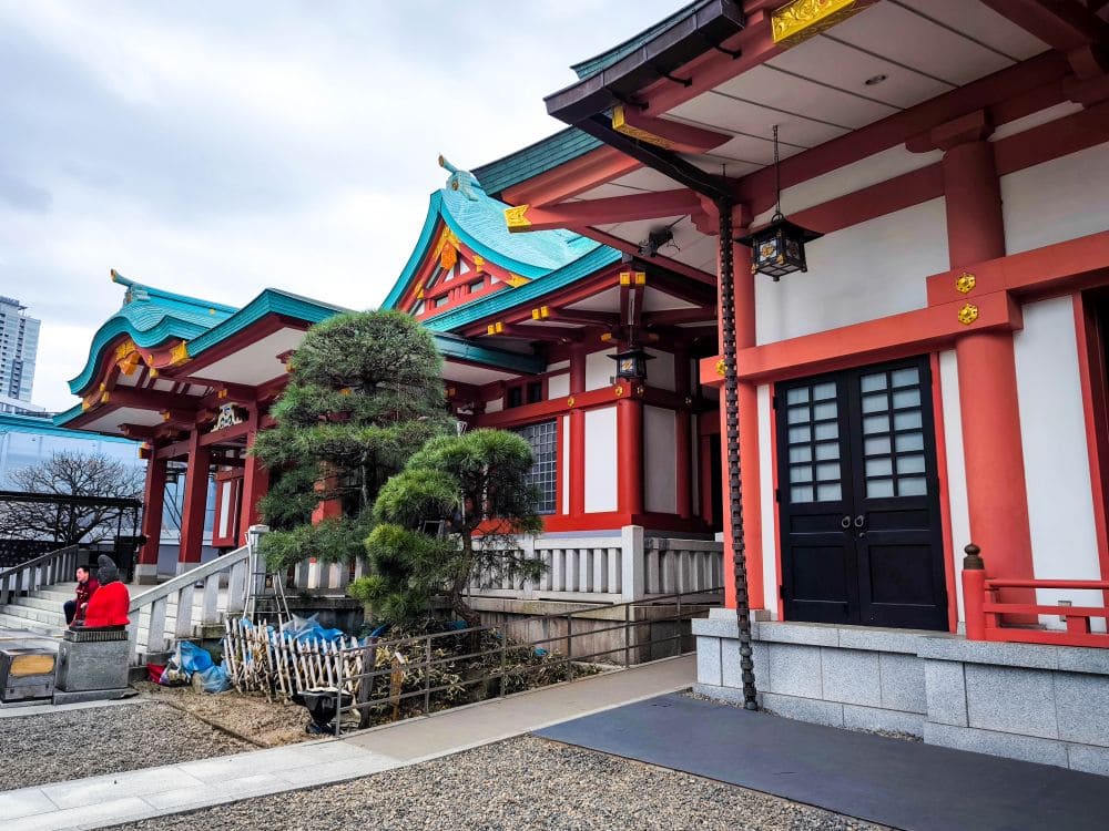 Ein traditionelles japanisches Gebäude in Hie Schrein besticht durch leuchtende Rot- und Blaugrüntöne und kunstvolle architektonische Details. Ein kleiner Baum ziert den Innenhof, und eine Person in Rot sitzt auf einer Steinbank neben einem stufenförmigen Eingang.
