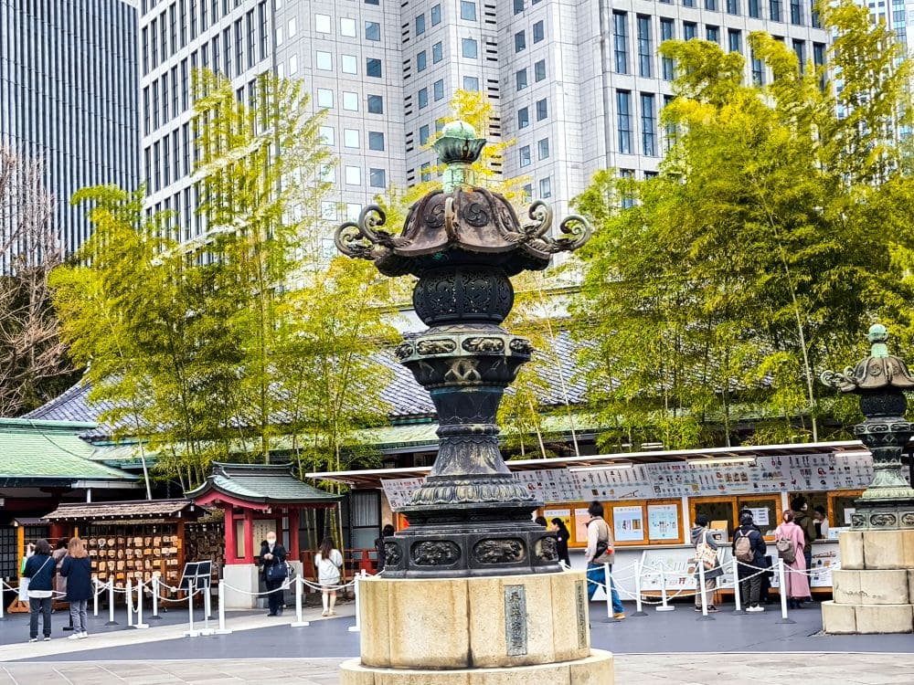 Eine traditionelle Bronzelaterne steht in einem Hof in der Nähe von Hie Schrein, umgeben von modernen Gebäuden. Hinter der Laterne sind Bambuspflanzen zu sehen. Menschen gehen umher und versammeln sich. Einige bleiben stehen, um sich die Schautafeln an einem nahegelegenen Stand anzusehen.