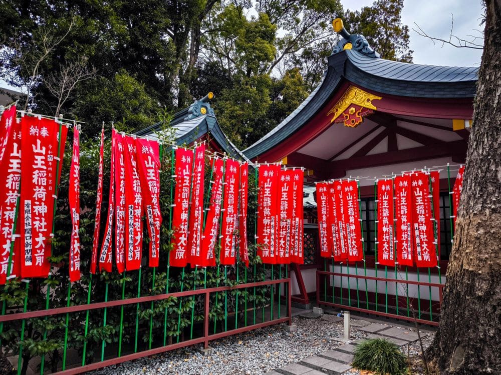 Bild des Hie Schreins, eines japanischen Schreins, geschmückt mit rot-weißen Bannern mit japanischen Schriftzeichen. Das Bauwerk besticht durch traditionelle Architektur mit kunstvollen Dachkonstruktionen. Die umliegenden Bäume bilden eine natürliche Kulisse, während sich Schotterwege durch die ruhige Gegend schlängeln.
