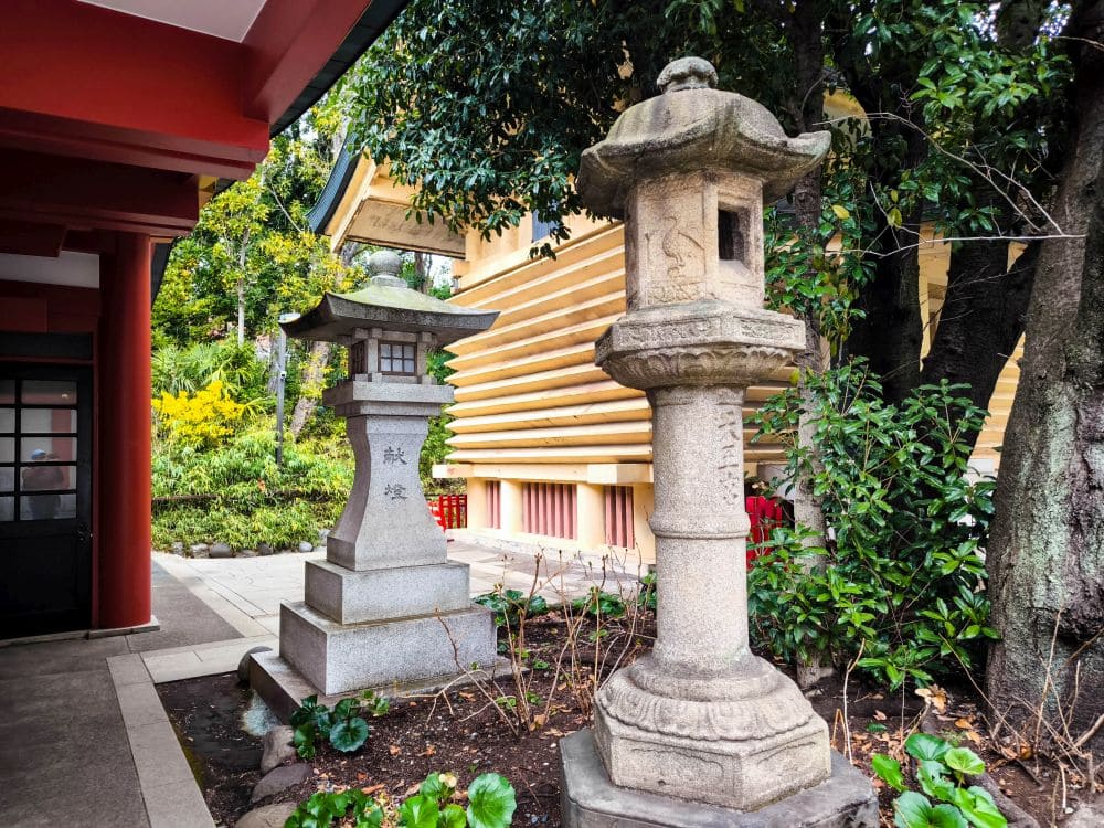 Ein malerischer Blick auf einen japanischen Garten in Hie Schrein mit zwei traditionellen Steinlaternen, üppigem Grün und einer Holzkonstruktion im Hintergrund. Der teilweise sichtbare Weg und die Gebäudeelemente unterstreichen die ruhige Schönheit traditioneller Architektur.