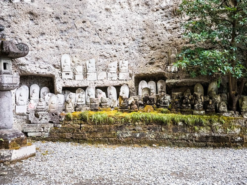 Eine Sammlung kleiner buddhistischer Steinstatuen und Schnitzereien im Ōya-ji-Tempel in Utsunomiya, die auf einem moosbewachsenen Felsvorsprung mit einem Baum in der Nähe und kiesigem Boden angeordnet sind - ein echter spiritueller Schatz.