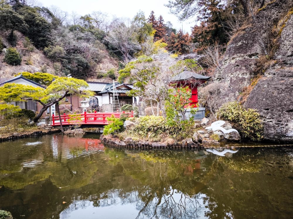 In einem friedlichen japanischen Garten am Ōya-ji-Tempel in Utsunomiya führt eine rote Brücke über einen Teich, der von Bäumen und Felsen umgeben ist. Ein traditionelles Gebäude und eine Steinlaterne, spirituelle Schätze, spiegeln sich im ruhigen Wasser.