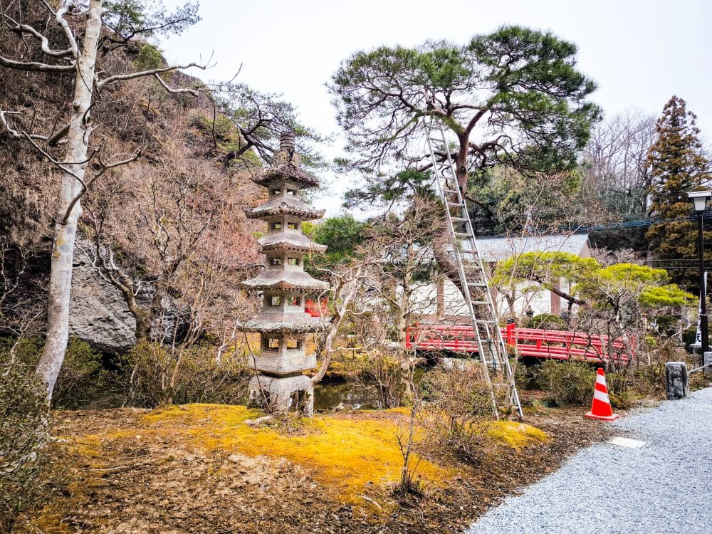 Ein japanischer Garten in Utsunomiya mit einer steinernen Pagodenlaterne, einer Metallleiter und einer roten Holzbrücke im Hintergrund bietet einen Blick auf einen spirituellen Schatz. Bäume, einige davon kahl verzweigt, und ein Verkehrskegel säumen den Kiesweg in der Nähe des Ōya-ji-Tempels.