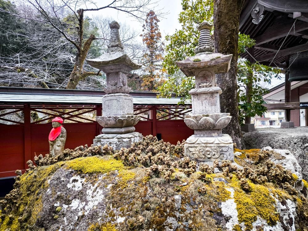 Zwei Steinlaternen und eine kleine Statue mit rotem Lätzchen ruhen auf einem moosbewachsenen Felsen im Ōya-ji-Tempelgarten in Utsunomiya, einem Ort, der als Geisterschatz verehrt wird. Kahle Bäume und ein roter Holzzaun rahmen die heitere Szene ein.