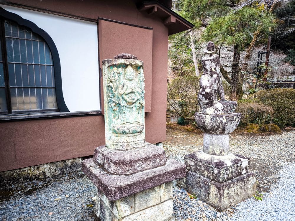 Zwei verwitterte Steinstatuen auf Sockeln stehen vor einem braunen Gebäude mit einem großen Fenster in Utsunomiya, umgeben von Büschen und Bäumen in einem Kieshof - ein Echo auf den Geisterschatz des historischen Ōya-ji-Tempels.