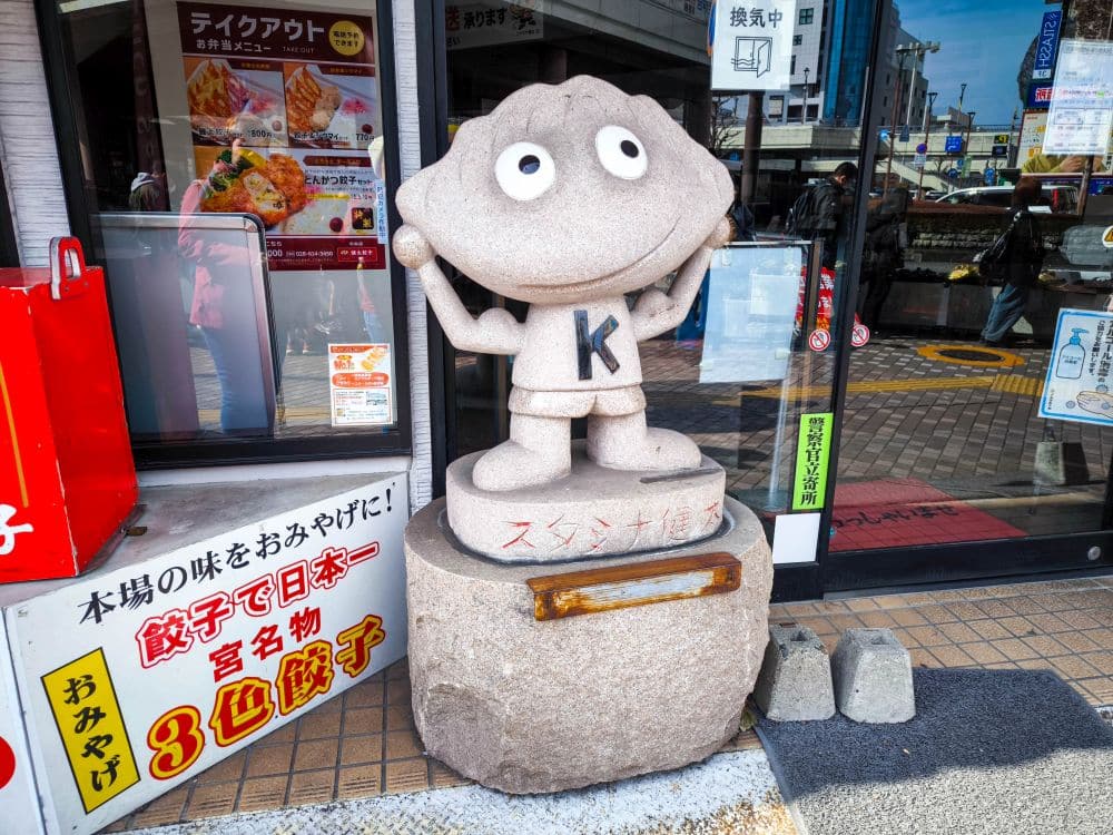 Eine steinerne Statue einer Zeichentrickfigur mit großen Augen, einem runden Kopf und einem Hemd mit K steht vor einem Geschäft in Utsunomiya, Japan, das für seine Gyoza bekannt ist. Im Hintergrund sind japanische Schilder und Gebäudereflexionen zu sehen.