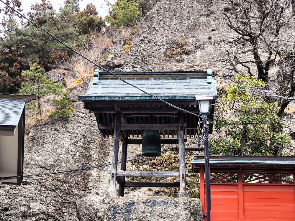 Ein traditioneller japanischer Glockenturm mit einer grünen Glocke steht neben einem roten Holzzaun am Ōya-ji-Tempel in Utsunomiya, einem spirituellen Schatz, der von felsigem Gelände und Bäumen umgeben ist und von Überlandleitungen durchzogen wird.
