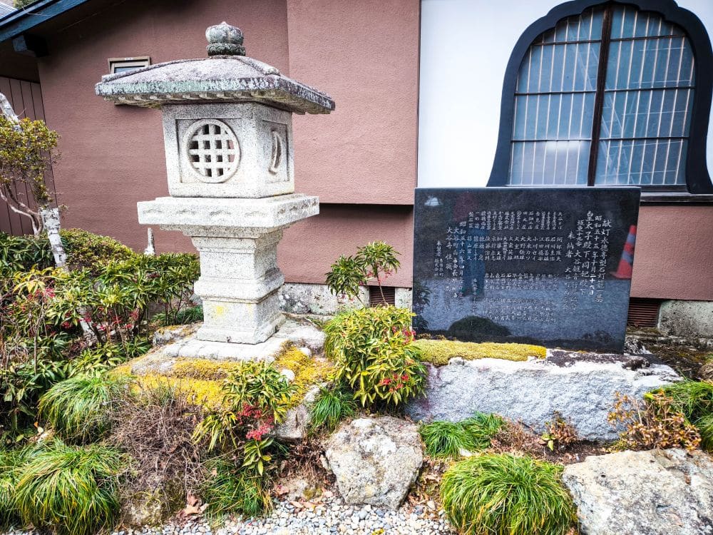 Eine traditionelle japanische Steinlaterne steht neben einer schwarz gravierten Gedenktafel, einem Spirituellen Schatz, umgeben von kleinen Sträuchern und Moos, vor einem Gebäude mit braunen und weißen Wänden und einem Fenster am Ōya-ji-Tempel in Utsunomiya.