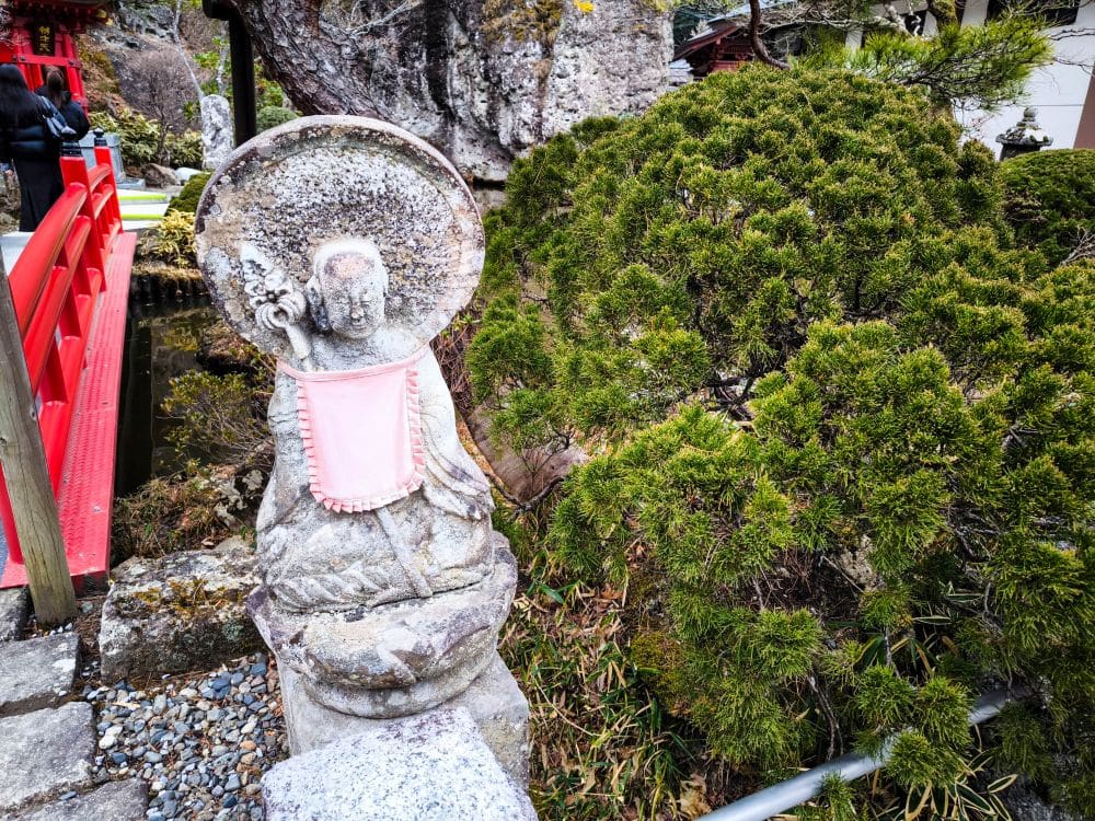 Eine verwitterte Steinstatue, die ein rosa Lätzchen trägt, steht neben grünen Sträuchern in der Nähe einer roten Holzbrücke in einer japanischen Gartenanlage, die an die spirituellen Schätze des historischen Ōya-ji-Tempels in Utsunomiya erinnert.