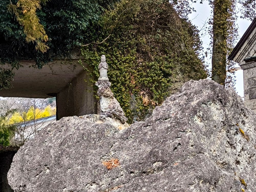 Am Ōya-ji-Tempel in Utsunomiya steht eine kleine Steinstatue auf einem großen, rauen Felsblock, umgeben von Grün und Ranken, und bietet einen Blick auf den Geisterschatz mit einem teilweise sichtbaren Gebäude im Hintergrund.