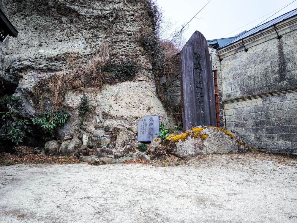 Ein Steindenkmal mit japanischen Inschriften steht neben einer Felswand und einem kleineren gravierten Stein, umgeben von Sträuchern und Moos in der Außenanlage des Ōya-ji-Tempels, Utsunomiya - ein wahrer spiritueller Schatz.