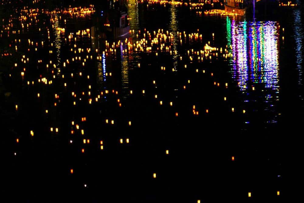 Hunderte von kleinen leuchtenden Laternen schwimmen nachts auf dem dunklen Wasser, reflektieren die bunten Lichter der Stadt und schaffen eine heitere, festliche Atmosphäre, die an das japanische Fest Obon zu Ehren der Ahnen erinnert.