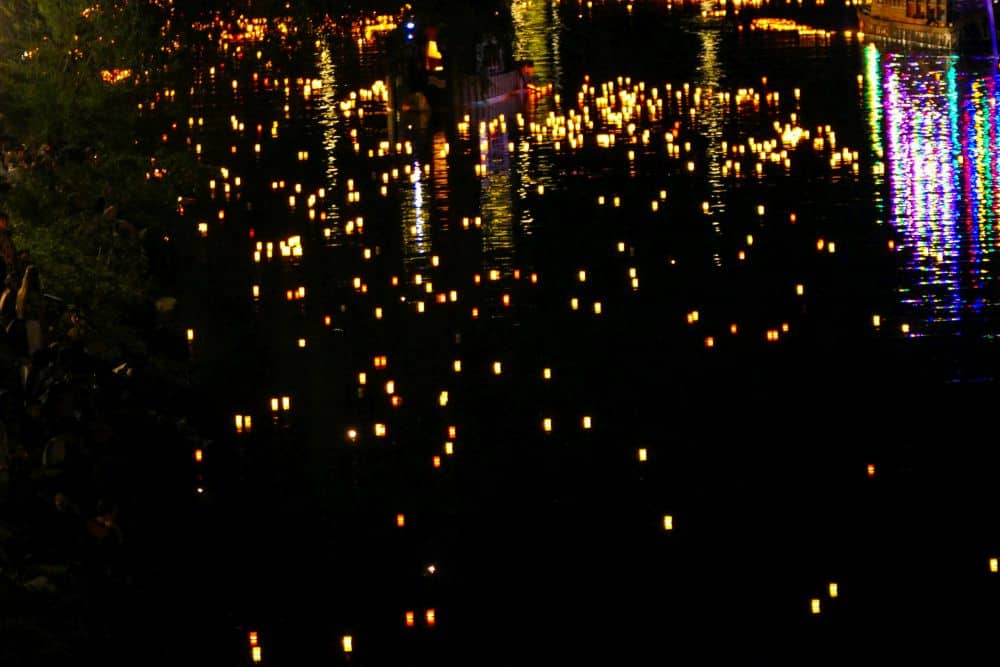 Hunderte von leuchtenden Laternen treiben nachts während Obon, dem japanischen Fest zu Ehren von Ahnen, auf einem dunklen Fluss. Ihre Lichter werden vom Ufer reflektiert und schaffen eine friedliche, magische Szene, während sich die Menschen am Flussufer versammeln, um zuzusehen.