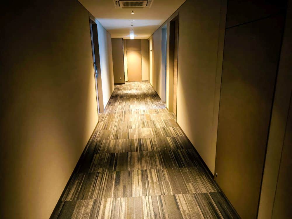 Ein schwach beleuchteter Flur mit gemustertem Teppich, beigen Wänden und geschlossenen Türen auf beiden Seiten erweckt die geheimnisvolle Atmosphäre einer Tobu-Einrichtung. Das Licht der Deckenleuchten erhellt sanft das Ende des Flurs.