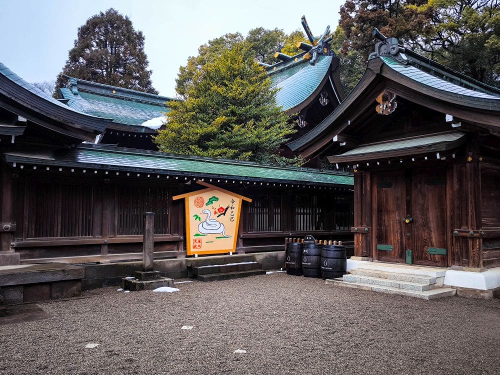 Ein traditioneller Hikawa-Schrein mit dunklen Holzgebäuden und grünen Ziegeldächern, mit einer farbenfrohen Ema-Tafel mit einer weißen Schlangenillustration und mehreren schwarzen Fässern am Eingang.
