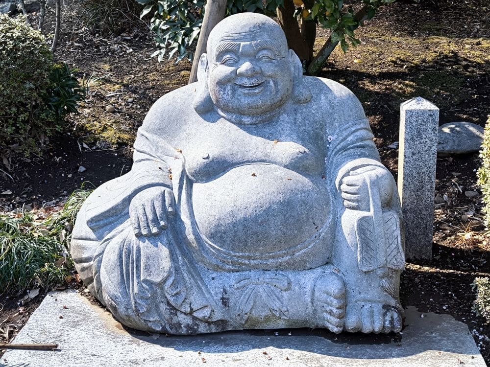 Eine Steinstatue eines lächelnden, sitzenden Buddhas mit rundem Bauch, der in einer Hand einen Fächer hält, steht im Freien zwischen Grünpflanzen und einer rechteckigen Steinmarkierung am Hosenji-Tempel in Hino und spiegelt die lokale Geschichte wider.
