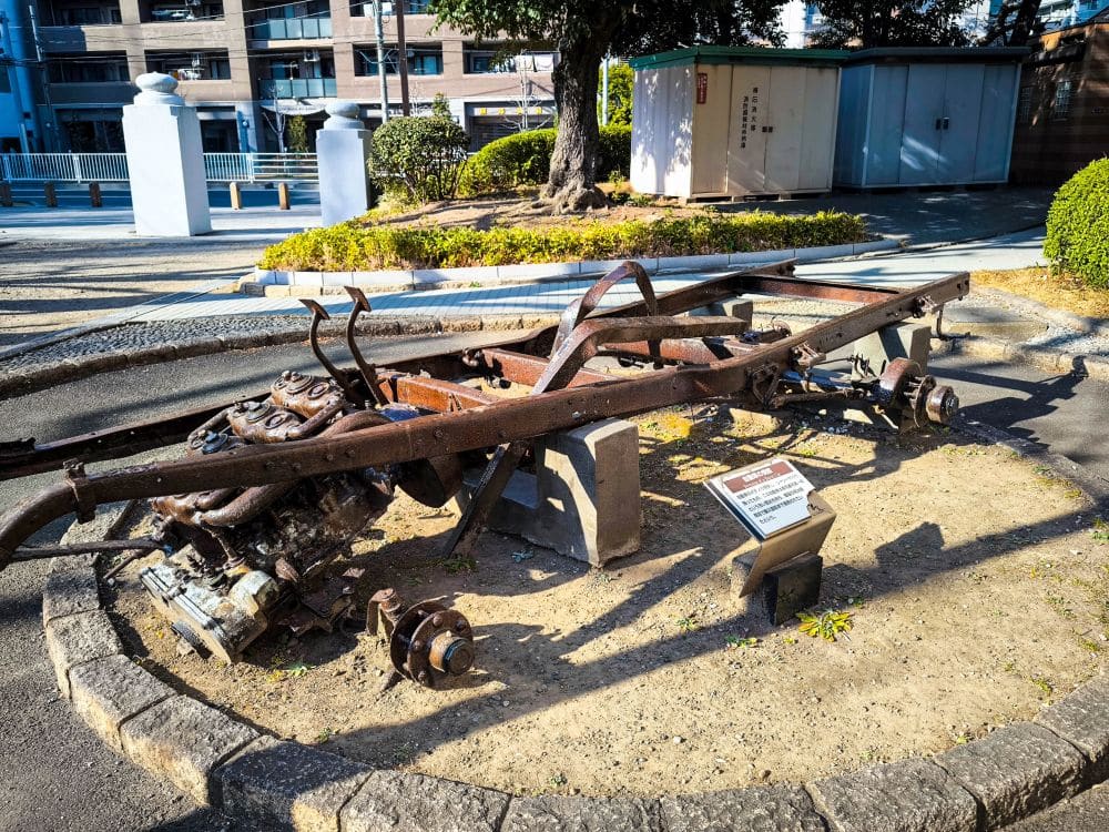 Ein verrostetes, skelettiertes Autochassis steht im Freien auf einem kreisrunden Fleck Erde, davor ein Metallschild, das auf die Geschichte des Memorial Museums hinweist. Im Hintergrund sind Gebäude, Sträucher und ein weißer Zaun zu sehen. Earthquake
