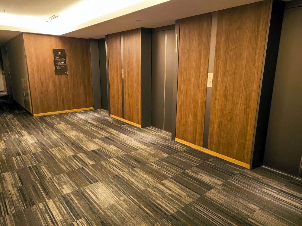 Ein moderner Tobu-Flur mit gemustertem Teppichboden, mit drei geschlossenen hölzernen Aufzugstüren und einem Wandschild auf der linken Seite; der Bereich ist durch Deckeneinbauleuchten gut beleuchtet.