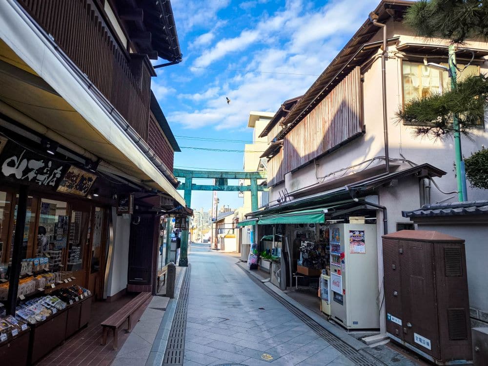 Eine schmale Straße in Enoshima, Japan, ist auf beiden Seiten von traditionellen Geschäften gesäumt und führt zu einem entfernten Torii-Tor unter einem blauen Himmel mit vereinzelten Wolken. Ein einzelner Vogel fliegt über der Szene.