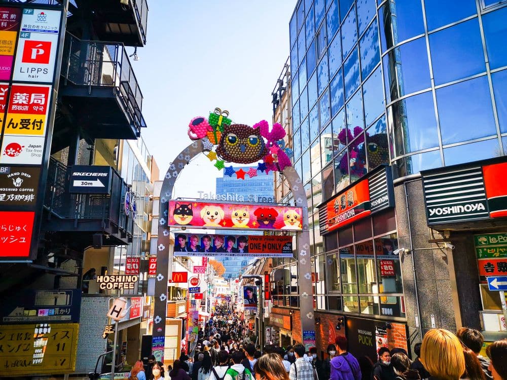 Der farbenfrohe Eingang zur Takeshita Street in Harajuku, Tokio, zeichnet sich durch einen großen dekorativen Bogen, lebhafte Menschenmengen, Geschäfte und leuchtende Schilder aus, die beide Seiten dieser belebten und ikonischen Straße säumen.