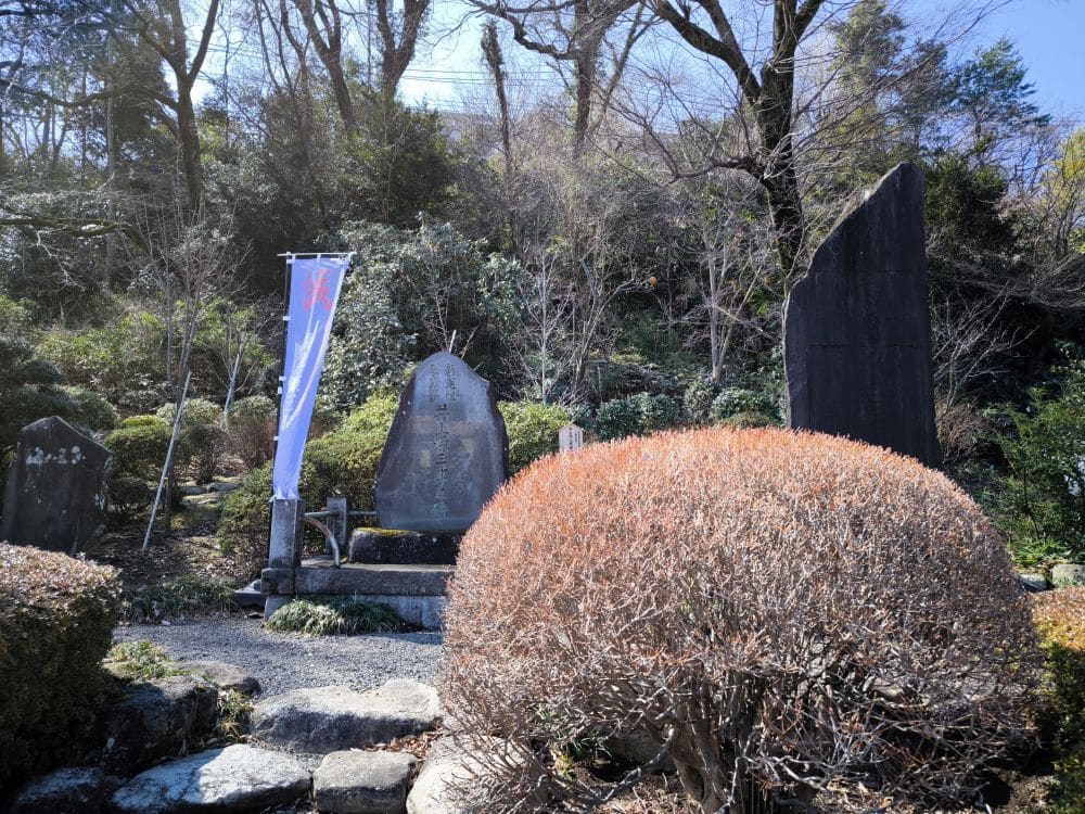Eine ruhige Szene im Freien am Hosenji-Tempel in Hino, mit Steindenkmälern und einer großen Fahne mit japanischen Schriftzeichen, umgeben von gepflegten Büschen und Bäumen. Ein Schotterweg und Felsen führen die Besucher durch diese historische Umgebung.