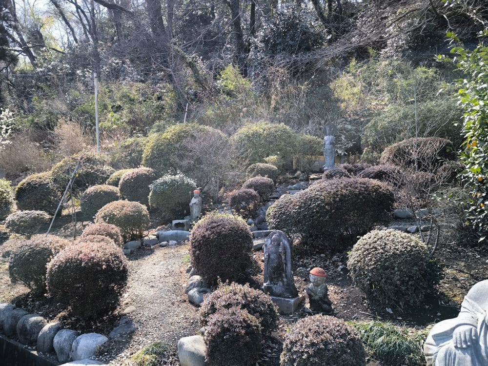 Ein ruhiger Garten am Hosenji-Tempel in Hino, mit ordentlich gestutzten Büschen, Statuen und kleinen Steintafeln zwischen dem Laub. Das Sonnenlicht fällt durch die Bäume und spiegelt die reiche Geschichte und friedliche Atmosphäre des Tempels wider.