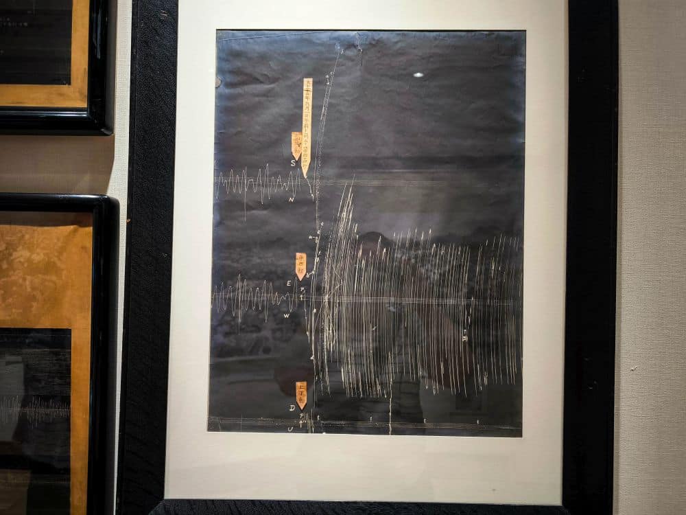 Ein gerahmtes Stück schwarzes Papier, das einen Seismographen des Großen Kanto-Erdbebens zeigt, mit gezackten weißen Linien, die die seismische Aktivität anzeigen, und kleinen gelben Etiketten, die an Teilen des Papiers angebracht sind, wie in einem Memorial Museum zu sehen.