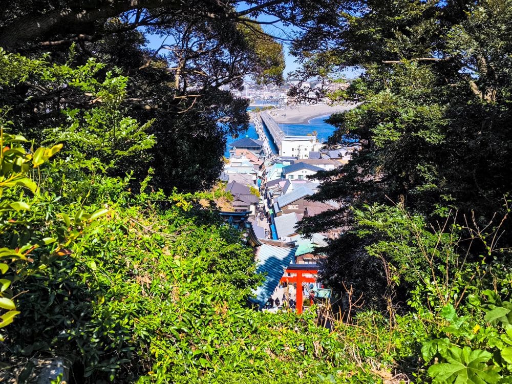 Blick durch dichte grüne Bäume auf die Küstenstadt Hetsunomiya mit ihren Dächern, einem roten Torii-Tor und einer langen Brücke, die sich unter einem strahlenden, sonnigen Himmel über das blaue Wasser erstreckt.