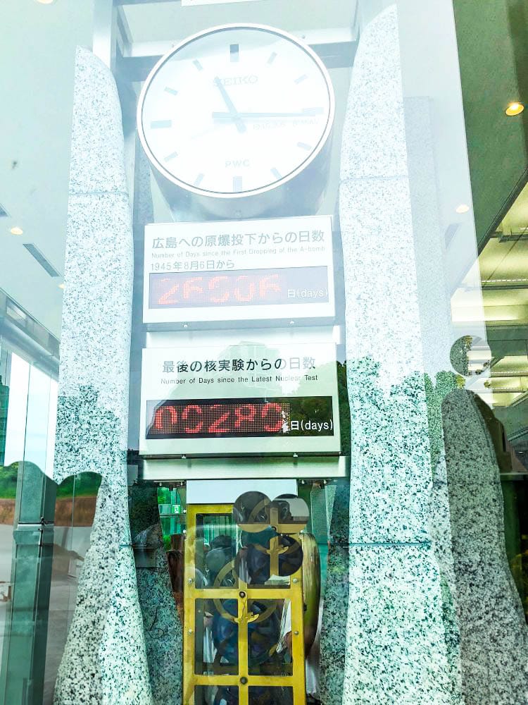 Eine große Uhr über digitalen Zählern im Hiroshima Peace Memorial Museum zeigt die Tage seit dem Atombombenabwurf und dem letzten Atomtest an und symbolisiert einen Aufruf zum Frieden. Die Spiegelungen der Besucher sind im Glas sichtbar.
