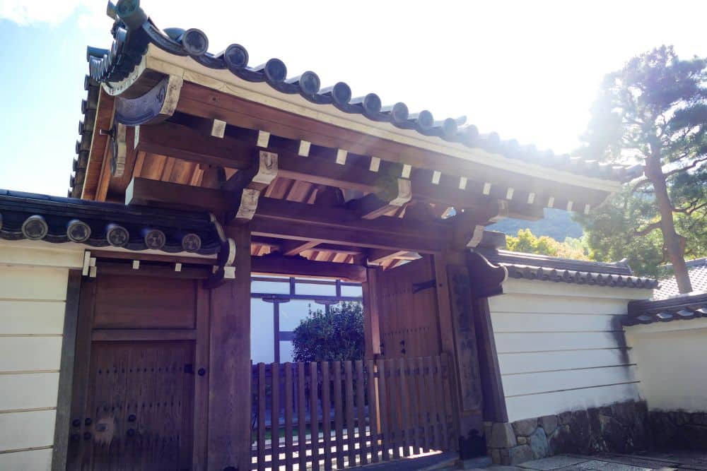Ein traditionelles japanisches hölzernes Tempeltor mit einem Ziegeldach steht offen und ist an weißen Wänden befestigt. Das von oben einfallende Sonnenlicht beleuchtet das Tor und wirft Schatten auf den Boden. Im Hintergrund sind Bäume und Hügel zu sehen.