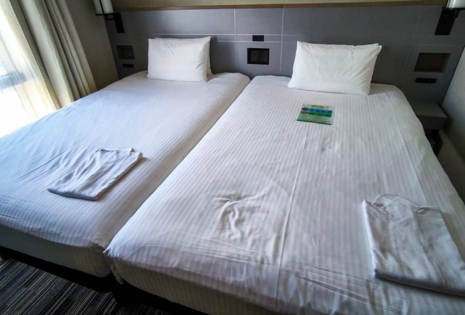 Zwei Einzelbetten mit weißer Bettwäsche und Kissen sind in diesem Tobu-Hotelzimmer nebeneinander aufgestellt. Auf jedem Bett des AirBNB liegt ein gefalteter weißer Bademantel, und auf dem rechten Bett liegt eine kleine Broschüre. Durch ein Fenster auf der linken Seite fällt sanftes Licht ein.