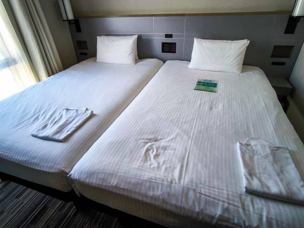 Zwei sauber gemachte Einzelbetten mit weißer Bettwäsche stehen nebeneinander in einem gemütlichen Zimmer des Asakusa Tobu Hotels. Auf jedem Bett liegt ein Handtuch, und auf dem rechten Bett liegt eine Broschüre. Durch ein Fenster auf der linken Seite strömt Licht herein.