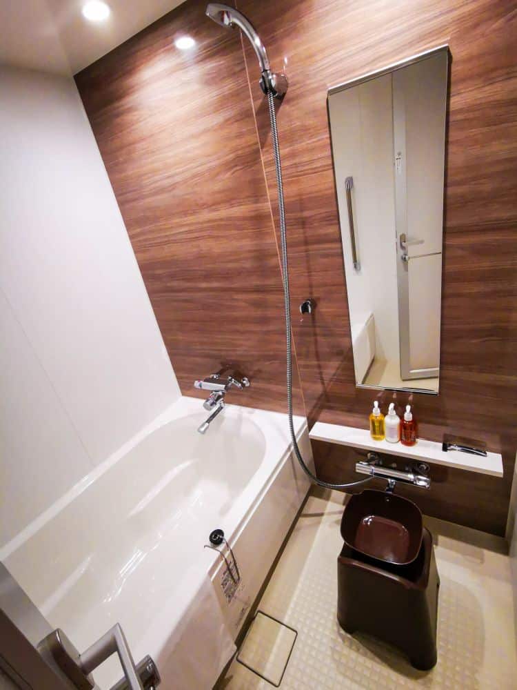 Ein modernes Badezimmer im japanischen Tobu-Stil mit einer tiefen Badewanne, einer Handbrause, einem hohen Spiegel, einem kleinen Hocker, einem Waschbecken und ordentlich angeordneten Toilettenartikeln auf einem Regal vor einer holzgetäfelten Wand.