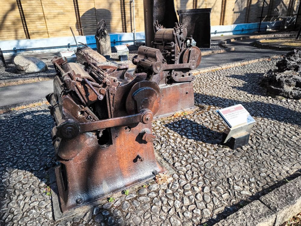 Eine große, verrostete Industriemaschine steht im Freien auf einem Kopfsteinpflaster mit einem Informationsschild in der Nähe. Sie ist Teil des Gedenkmuseums, das der Geschichte des großen Kanto-Erdbebens gewidmet ist; im Hintergrund sind weitere Maschinen und ein Bambuszaun zu sehen.