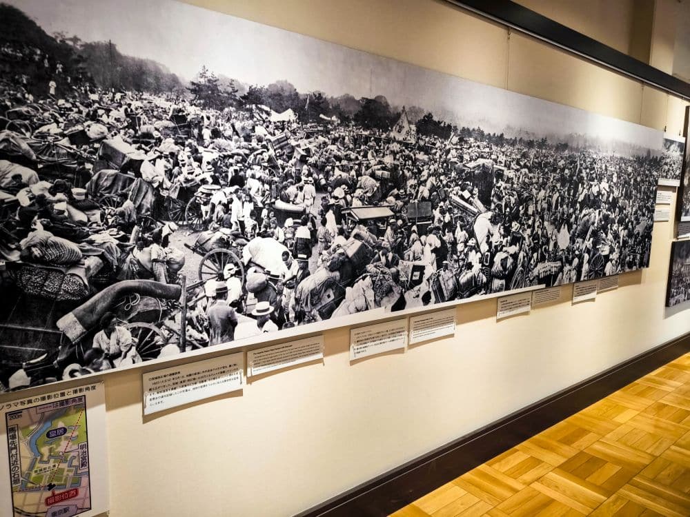 Eine Schwarz-Weiß-Wandmalerei im Memorial Museum zeigt eine dichte Menschenmenge, die ihre Habseligkeiten trägt, und erinnert an die Massenwanderung nach dem großen Kanto-Erdbeben. Darunter befinden sich Informationstafeln und ein Lageplan, die die Verbindung der Besucher zur Geschichte vertiefen.