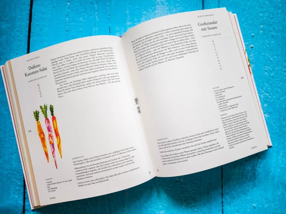 Auf einer hellblauen Holzplatte liegt ein aufgeschlagenes Kochbuch mit Rezepten in deutscher Sprache, mit Abbildungen von bunten Karotten auf der linken Seite und vor allem Text - voller Geschichte - auf den beiden Seiten.
